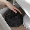 A hand is placing a black collapsible mesh laundry basket with side pockets into a white storage bin.