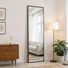 A tall, rectangular full body wall mirror with a black frame reflecting a bright window and interior decor.