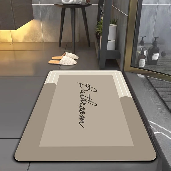A rectangular beige bathroom floor mat with the word Bathroom written in script on it, placed on a dark gray floor.