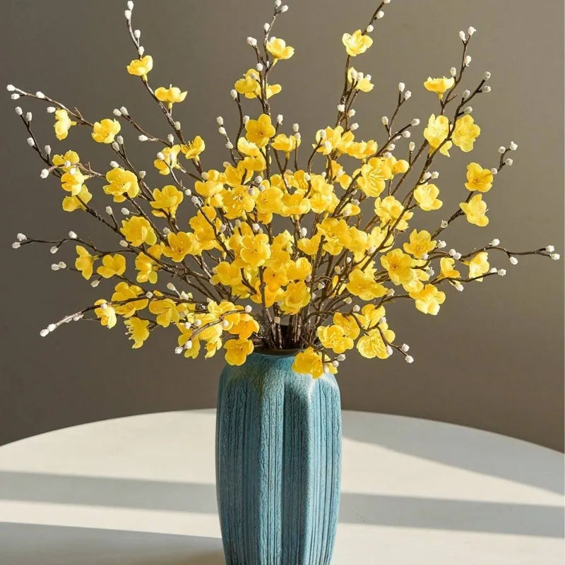 Eine Vase mit künstlichen gelben Pflaumenblütenzweigen auf einer weißen Oberfläche.