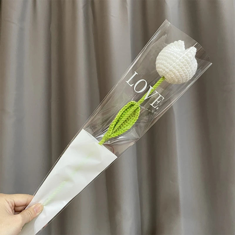 Une gaine en plastique transparent contenant une fleur de carnation blanche en coton et le mot AMOUR est tenue par une main c