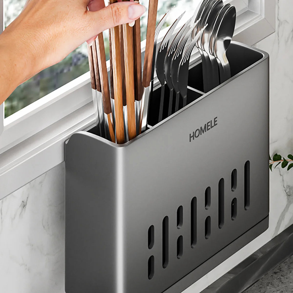 A hand is placing wooden and metal cutlery into a sleek, gray plastic storage rack with a drain base.
