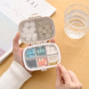 A hand holds an open 8-grid portable pill organizer with various colored pills and a clear glass cup on a wooden surface.