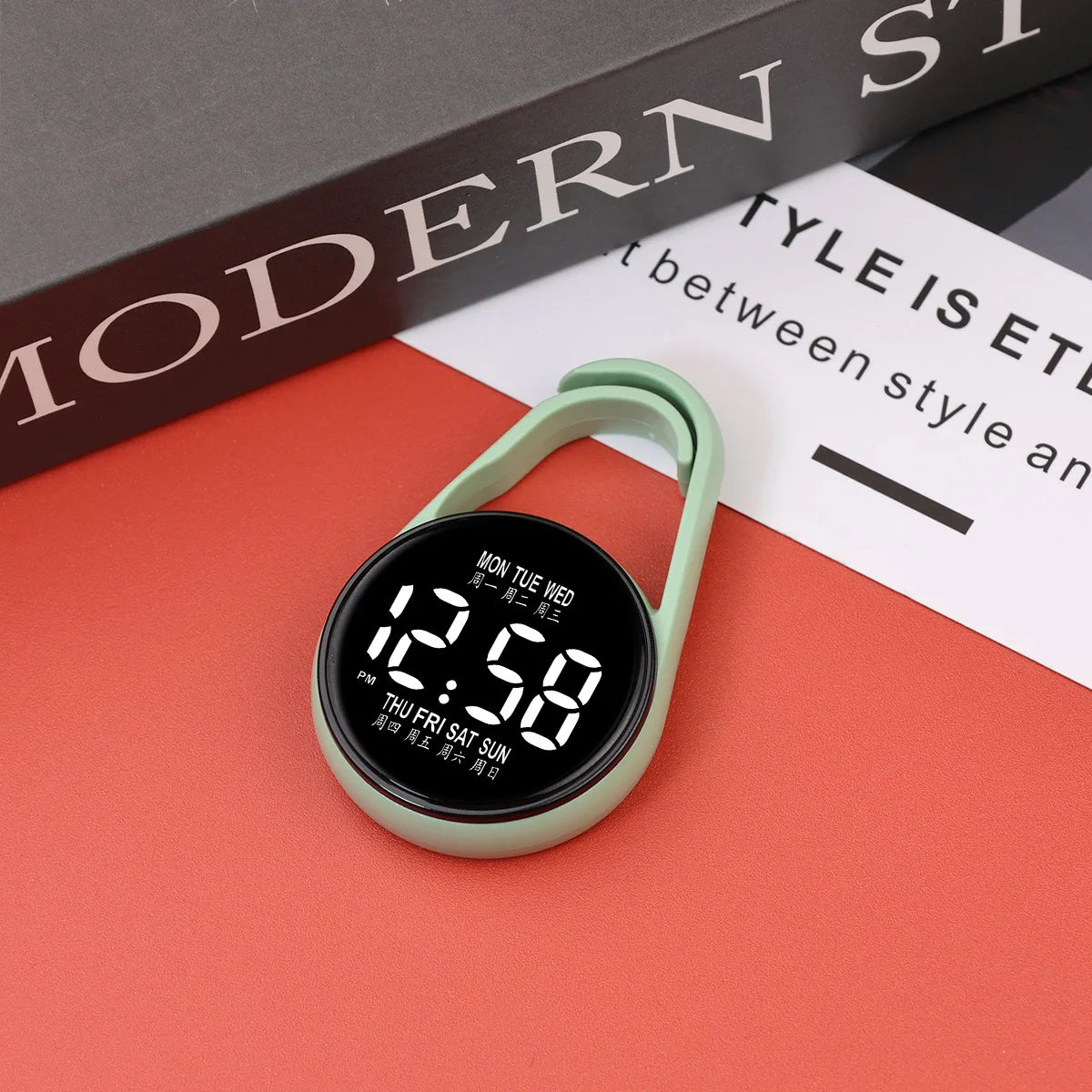 A digital LED hanging pocket watch with a light green strap and black face is displayed on an orange surface.