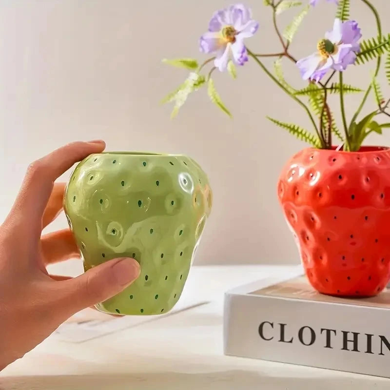Eine Hand hält eine hellgrüne, stachelbeerenförmige Harzblumenvase neben eine orange strukturierte Vase mit lila Blumen.