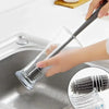A hand is using a silicone bottle brush with a long handle to clean inside a stainless steel sink.