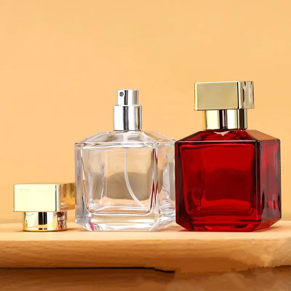 A clear glass perfume bottle with a silver cap stands next to a red glass perfume bottle with a gold cap on a wooden surface.