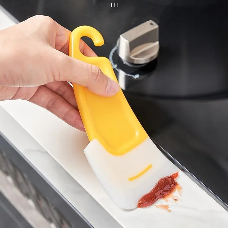 A hand is using a yellow silicone kitchen scraper to clean a dish on a white countertop.