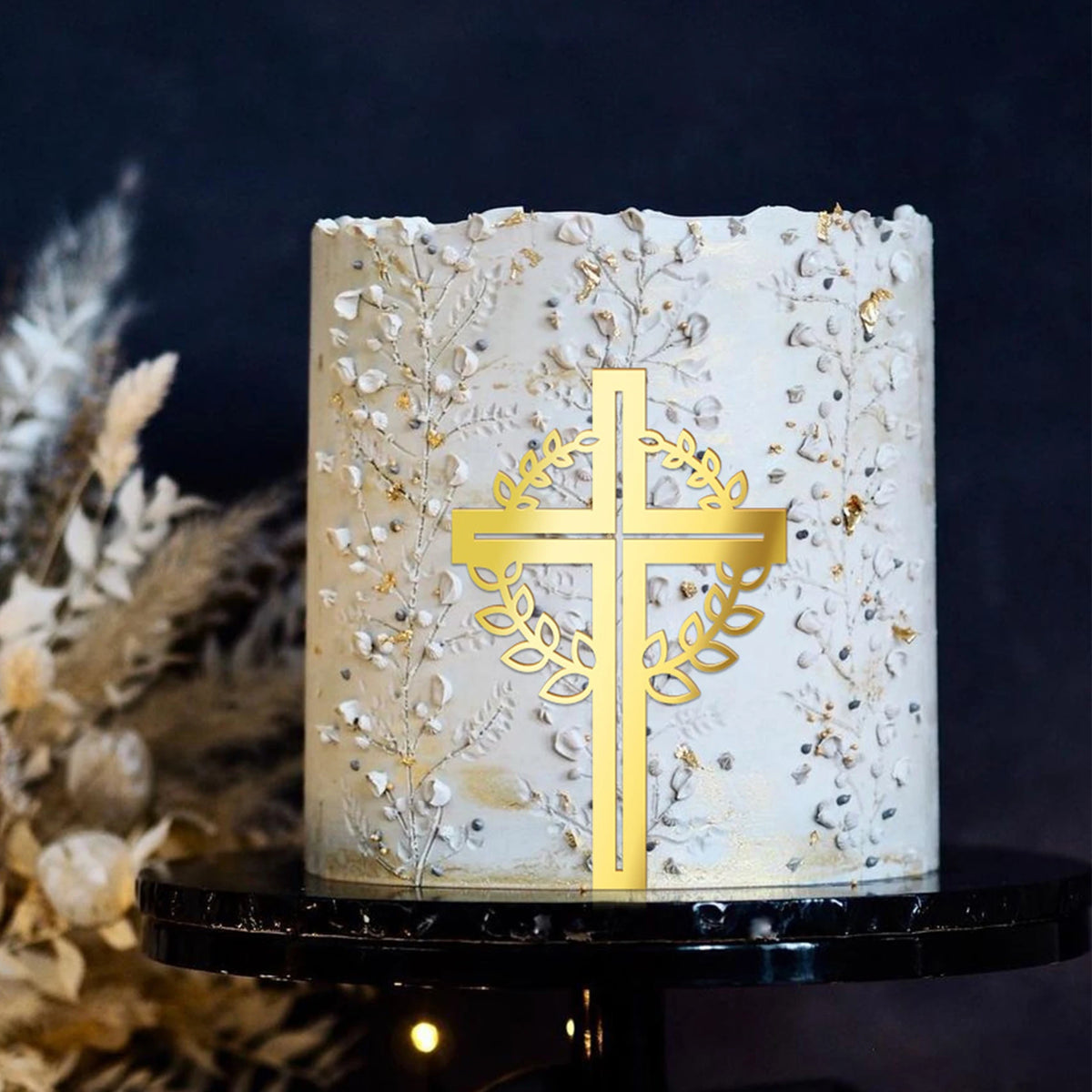 A cylindrical white cake decorated with gold acrylic cross toppers and dried floral accents.