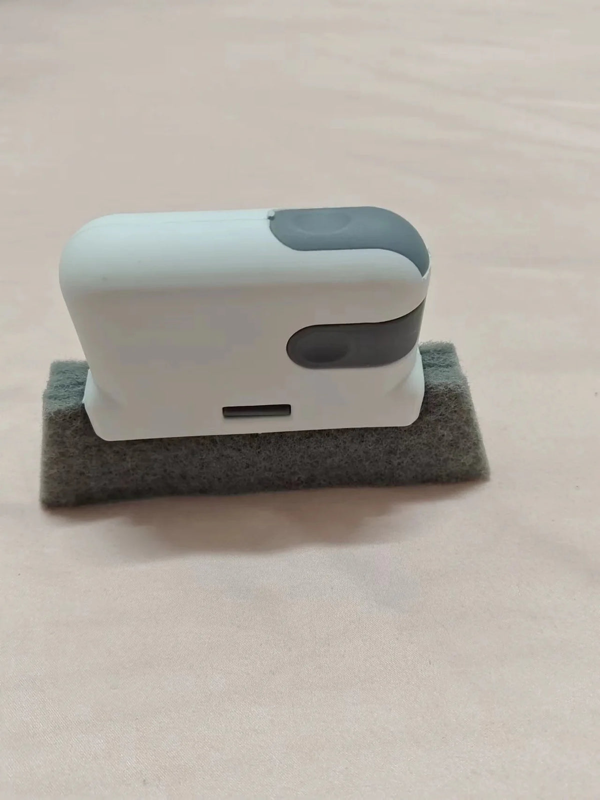 Una brocha de limpieza blanca y gris descansa sobre una pieza de espuma gris en una superficie rosa claro.