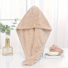 A beige microfiber hair drying wrap towel is displayed on a white surface next to a potted plant and a skincare product.