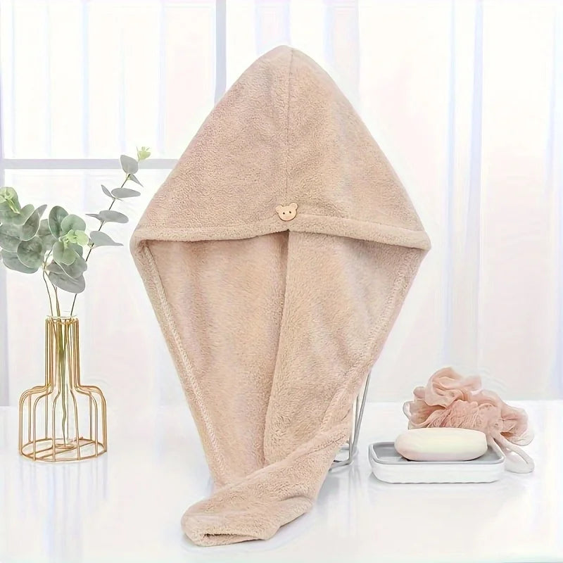 A beige microfiber hair drying wrap towel is displayed on a white surface next to a potted plant and a skincare product.