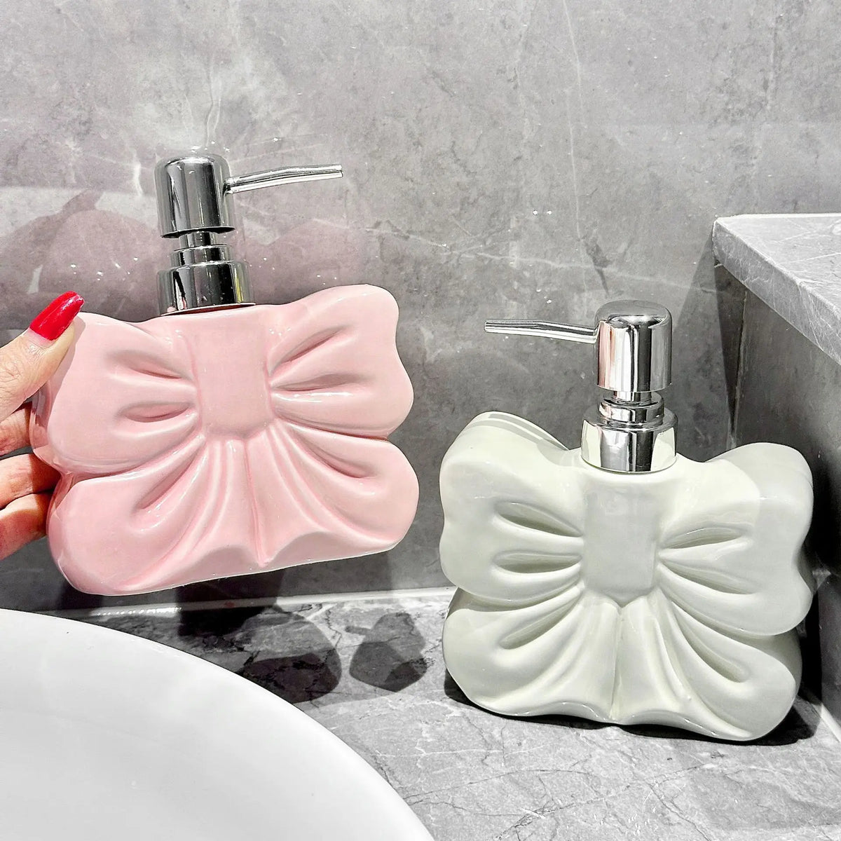 A pink ceramic pump dispenser bottle with a bowknot design is shown next to a white one on a marble surface.
