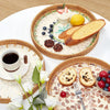 A round rattan serving tray with mother of pearl inlay holds sliced bread, fruit, and a cup.