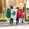 Two women walk past carrying a large Mexican flag in a sunny outdoor setting.