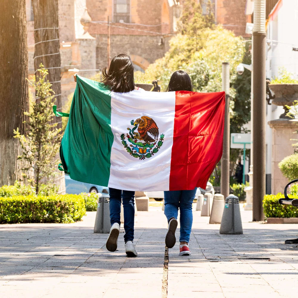 Two women walk past carrying a large Mexican flag in a sunny outdoor setting.