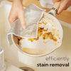 Hands are using a mesh cleaning cloth to remove stains from a round, light-colored cooking pan.