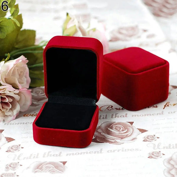 A square red velvet jewelry box with an open lid resting on a white floral background.