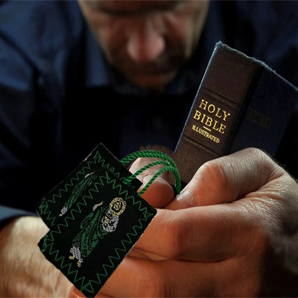 A hand holds a green scapular necklace set for Saint Jude and Virgen De Guadalupe against a dark background.