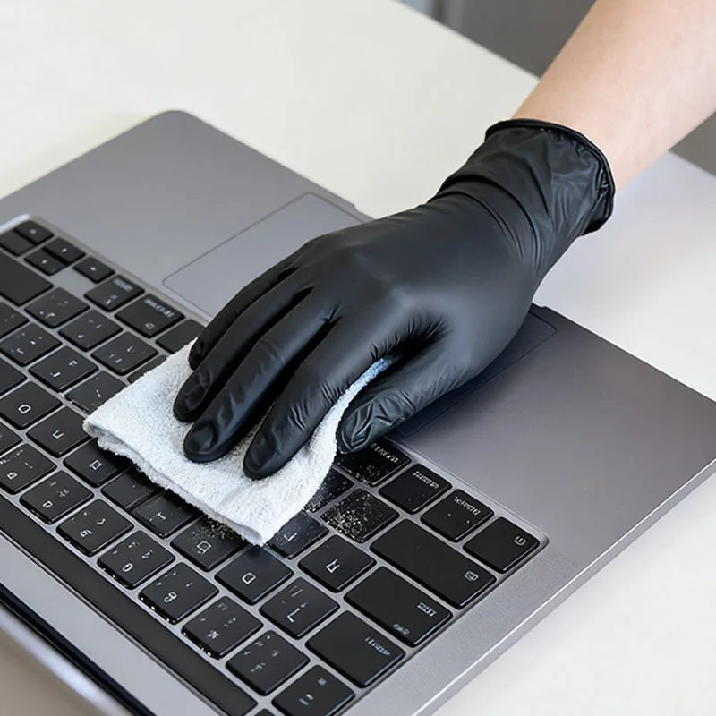 Una mano con un guante negro de nitrilo está limpiando el teclado de una laptop con un paño blanco.