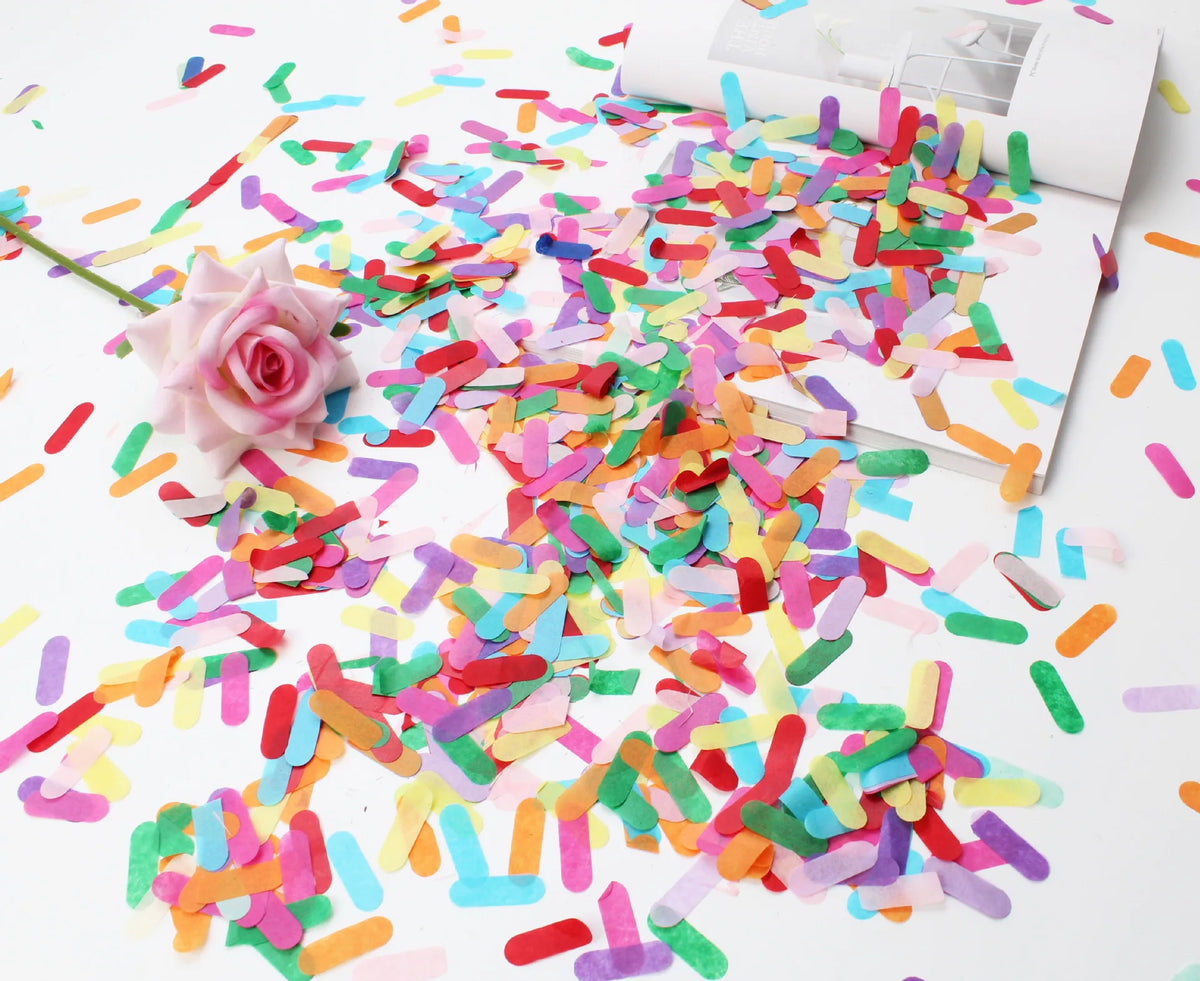 A colorful confetti decoration set scattered on a white surface next to a pink rose.