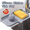A silicone kitchen sink tray holding a yellow baked item and a scouring pad being rinsed under water.