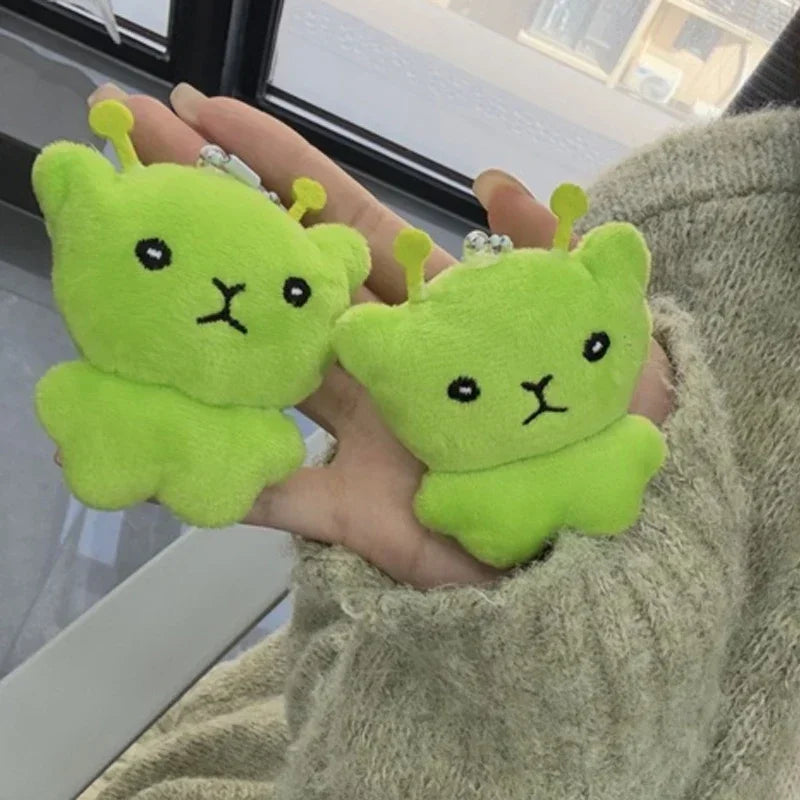 Two bright green plush cats are held in a person's hand against a window background.