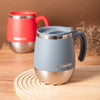 A light blue insulated stainless steel travel mug with a silver lid and a red mug in the background, resting on a wooden surf