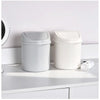 A white compact desktop trash can with a flip cover sitting on a white shelf.