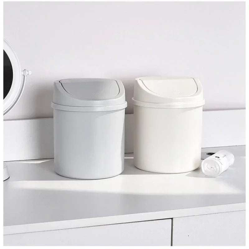 A white compact desktop trash can with a flip cover sitting on a white shelf.