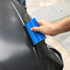 A hand uses a blue suede felt squeegee to smooth a black vinyl wrap or film on a surface.