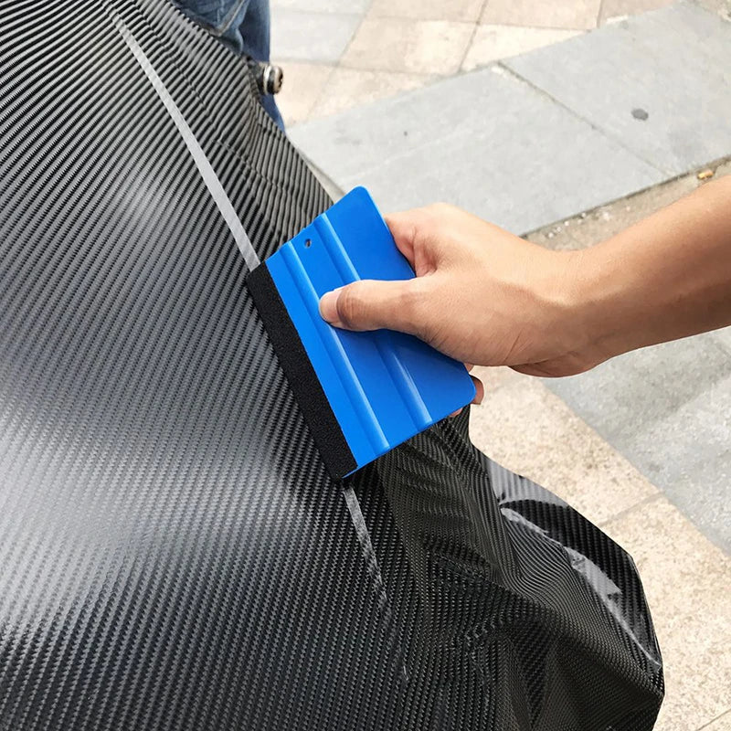 A hand uses a blue suede felt squeegee to smooth a black vinyl wrap or film on a surface.