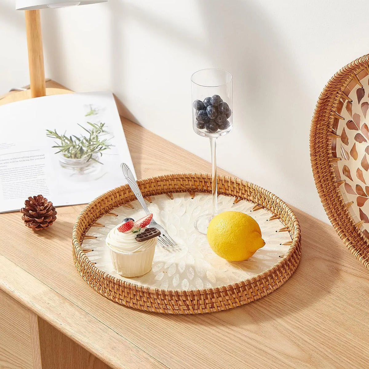 A round rattan serving tray with mother of pearl inlay holds a lemon, blueberries, and a small dessert.