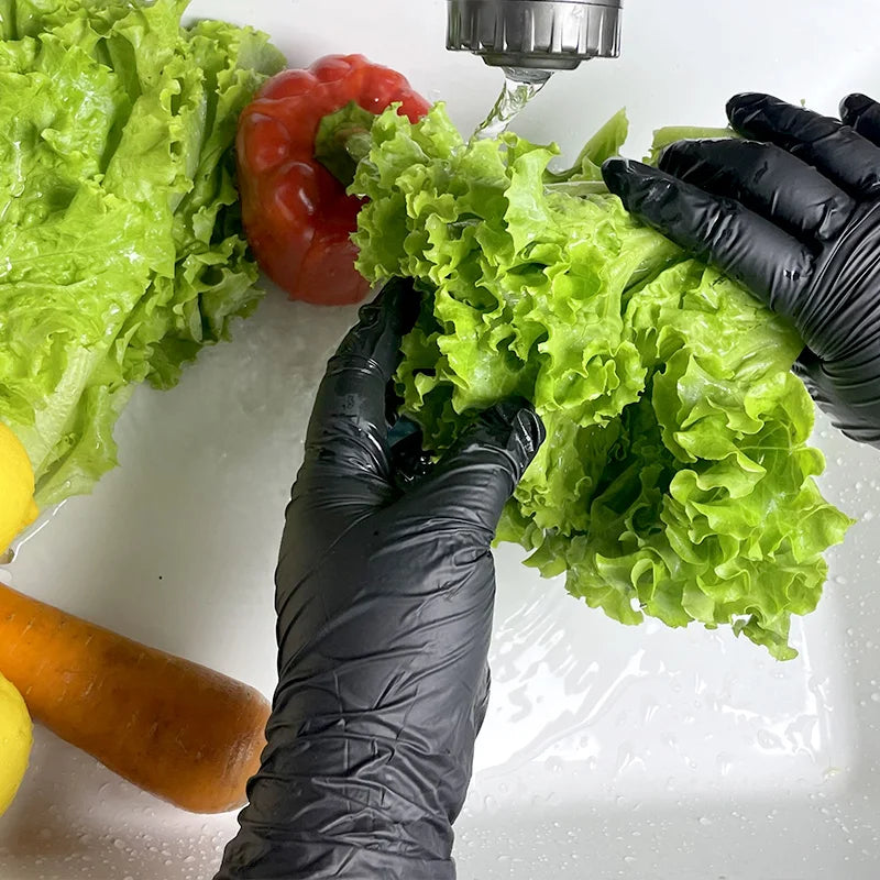 A hand wearing a black nitrile cleaning glove is holding a bunch of bright green lettuce and a red pepper on a white surface.