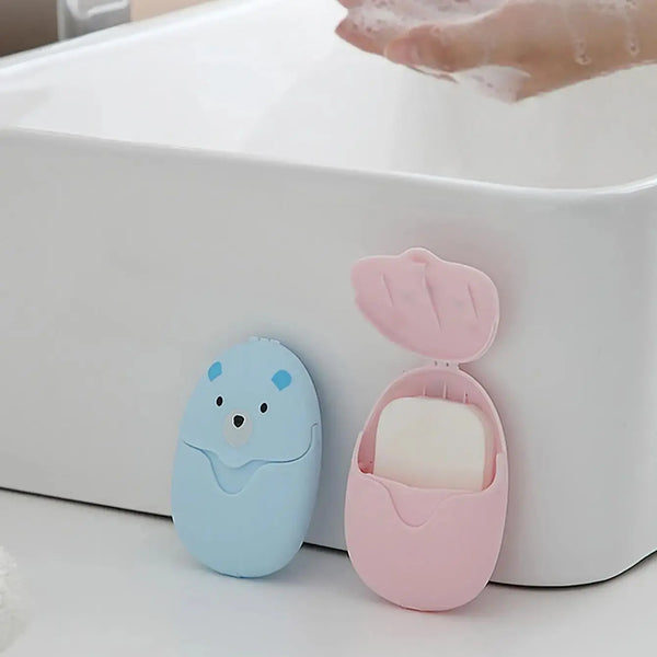 A pink portable paper soap dispenser with a lid open, showing a white soap bar inside, resting on a white tray.