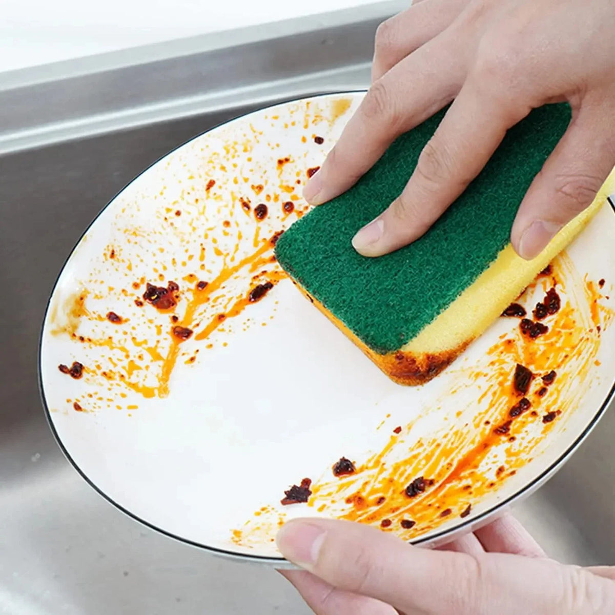 A hand is using a green and yellow magic cleaning sponge to wipe orange food residue off a white plate.