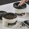 A hand is opening a white stainless steel vacuum sealed storage canister, revealing dark coffee beans inside.