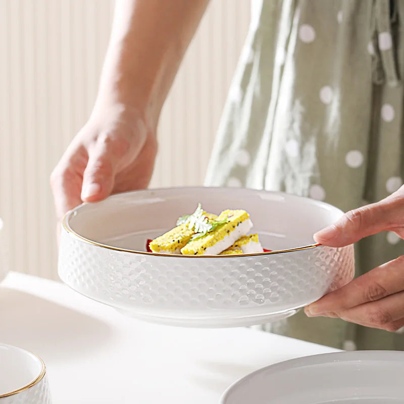 A hand is holding a white ceramic soup bowl filled with yellow and green food, set against a light background.
