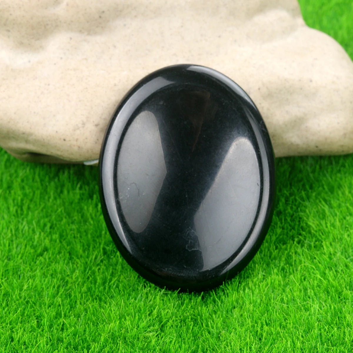 A smooth, black natural quartz thumb stone resting on vibrant green grass, held by a hand.