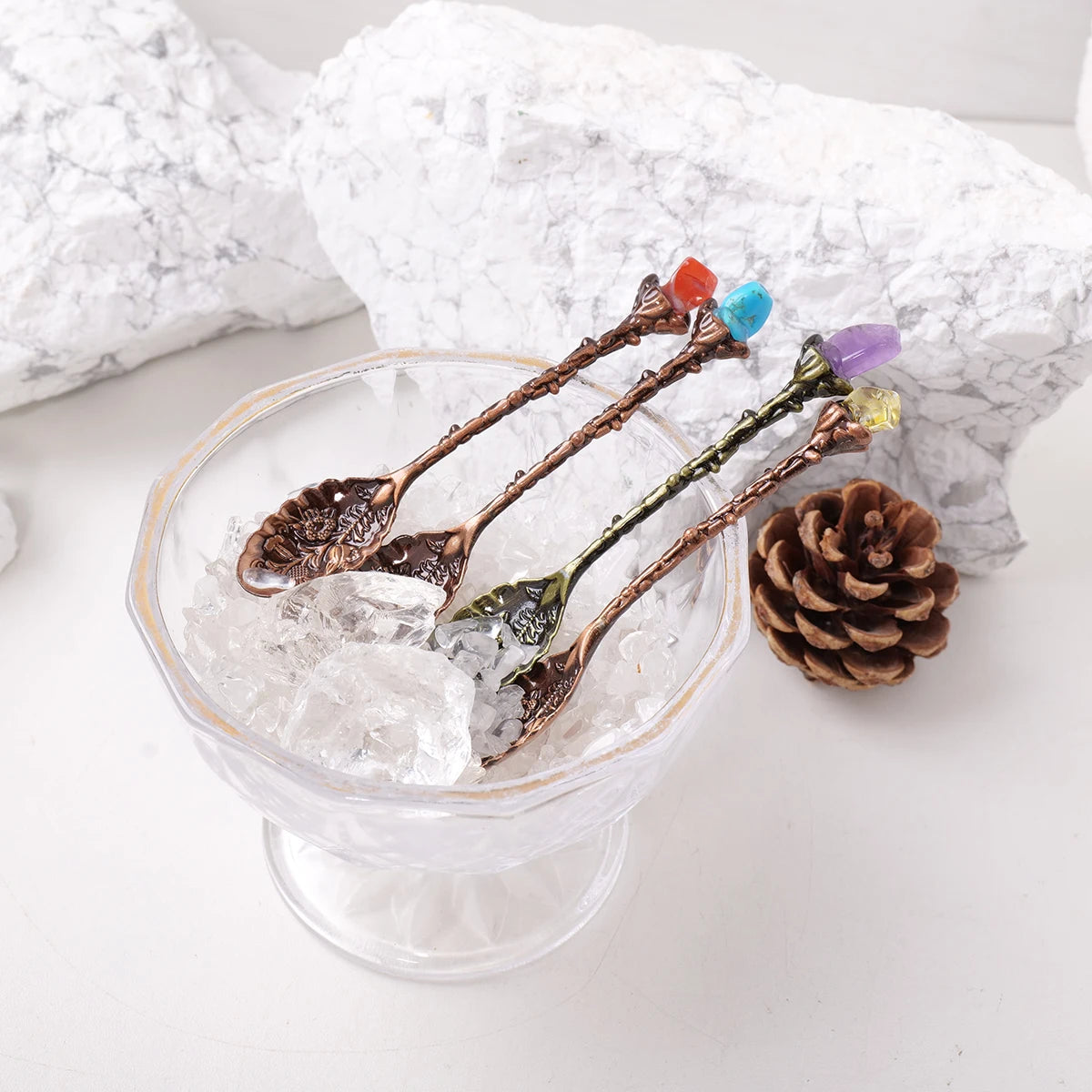 A glass filled with ice and several natural crystal spoons with colored crystals resting on a white surface.