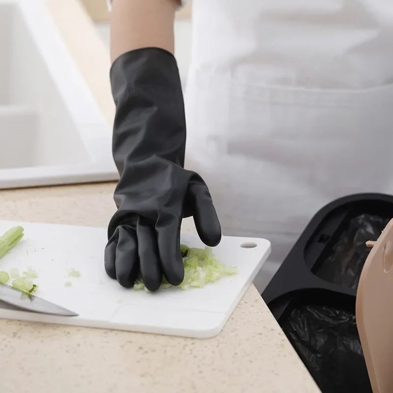 A hand wearing a black reusable rubber cleaning glove is holding chopped green herbs on a white cutting board.