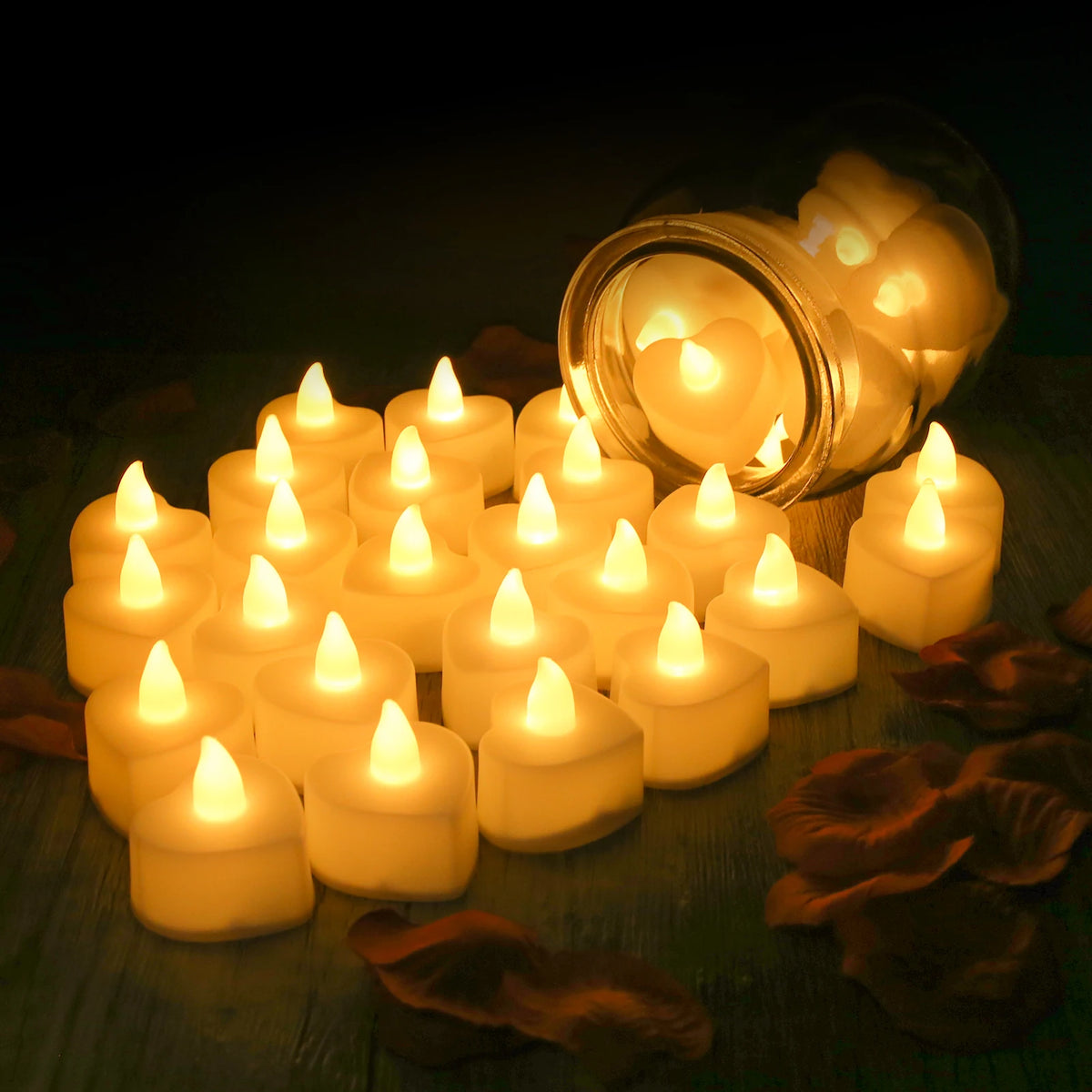 A glass jar filled with glowing LED heart tea lights resting on a wooden surface surrounded by artificial rose petals.