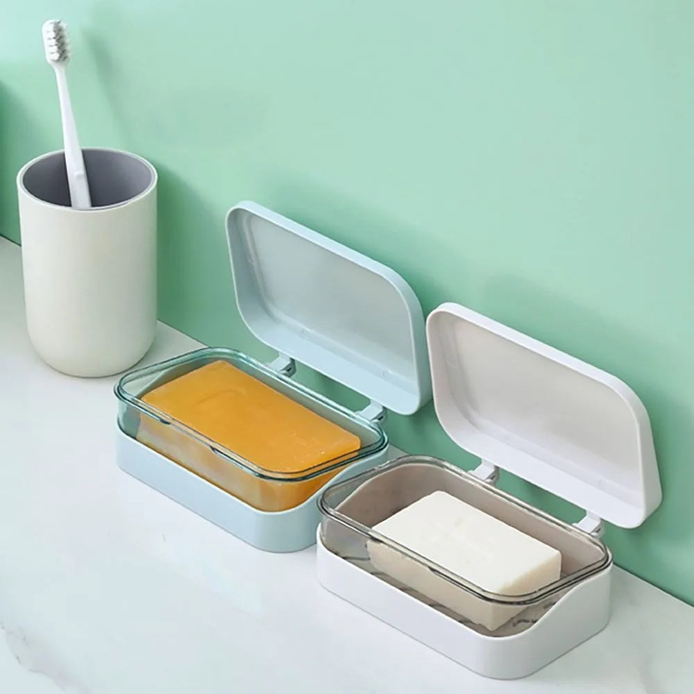 A white transparent drainage soap dish holds a bar of yellow soap next to a toothbrush on a light green wall.