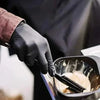A hand wearing a black nitrile glove is using a brush to apply product inside a black bowl.