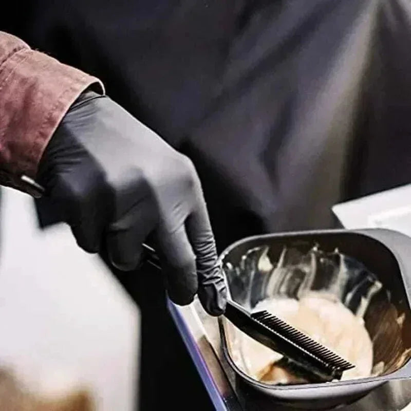 A hand wearing a black nitrile glove is using a brush to apply product inside a black bowl.