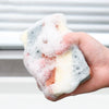 A hand holds a block of white and pink cleaning sponge covered in white foam.