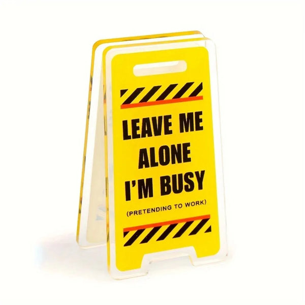 A yellow acrylic desk sign with black diagonal stripes that reads Leave Me Alone I'm Busy Pretending to Work