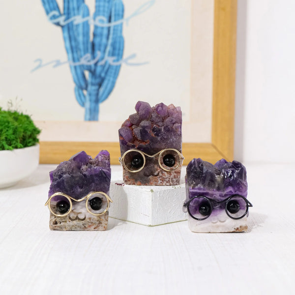 Three natural amethyst crystal wear glasses displayed on a white block against a blurred background of a blue cactus.