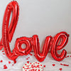 Rote herzförmige Ballonbriefe mit der Aufschrift LOVE liegen auf weißem Stoff mit verstreutem rotem Herzconfetti