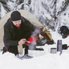 A person is cooking in a pot outdoors next to a tent in the snow with various aluminum cookware.